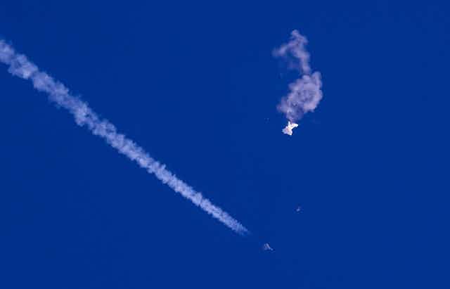 A photo showing wreckage of the Chinese balloon against the background of blue sky, with the vapour trail of a plane.