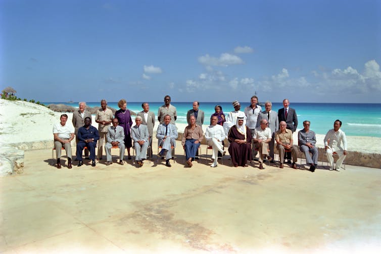 Two rows of people standing and sitting on a white beach with turquoise water in the background