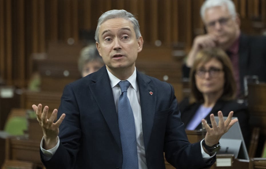 A white man with grey hair wearing a navy suit gestures with his hands while speaking