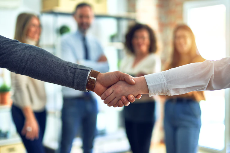 Two people shaking hands in front of a blurry background consisting of four people standing and smiling