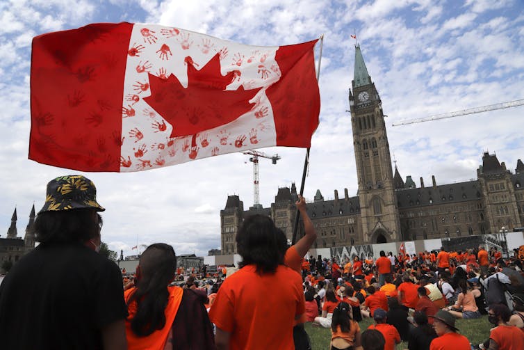 Protesters in orange T-shirts wave a Canadian flag with red handprints of children. The Peace Tower is in the background.