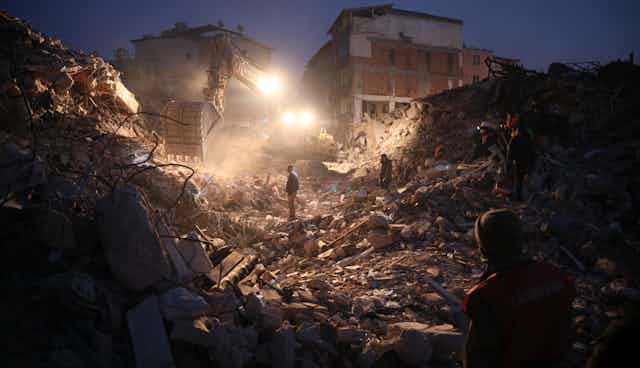People stand in the rubble of buildings illuminated by headlights.;.