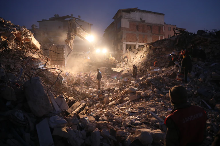 People stand in the rubble of buildings illuminated by headlights.;.