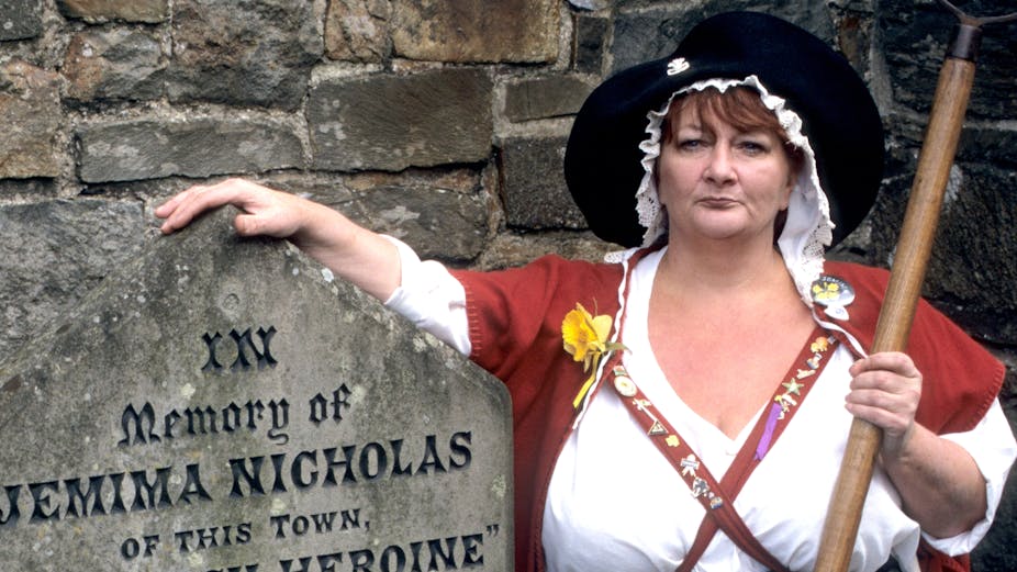 A woman wears the traditional Welsh dress of red wool and tall black hat. In her left hand she's carrying a pitchfork. Her right arm is draped over a gravestone saying "in memory of Jemima Nicholas"