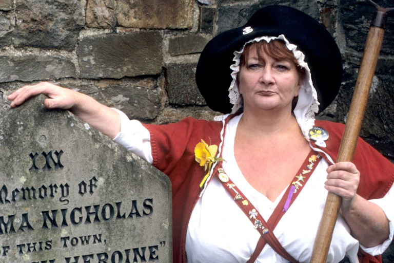 A woman wears the traditional Welsh dress of red wool and tall black hat. In her left hand she's carrying a pitchfork. Her right arm is draped over a gravestone saying "in memory of Jemima Nicholas"