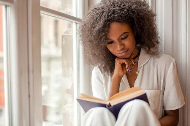A black woman sits on a windowsill reading a book. She wears a white shirt.