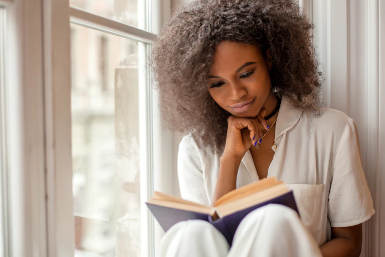 A black woman sits on a windowsill reading a book. She wears a white shirt.