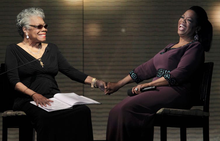 Two women seated on a stage hold hands.