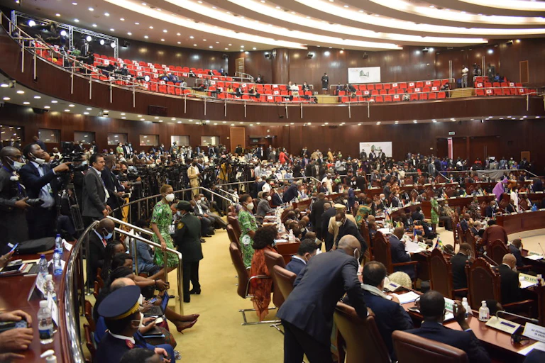 Men and women in a room, with groups of people seated or talking to each other, and another group of journalists pointing cameras towards the centre of the room