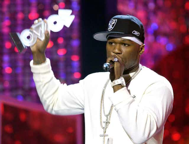50 Cent holds up his MOBO Award, wearing a white long sleeve shirt, black cap and two diamond crucifix chains.
