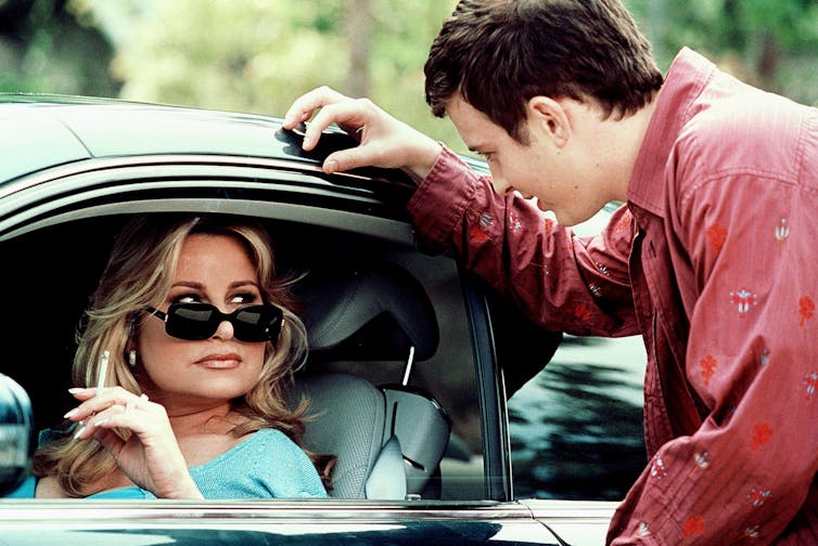 Jennifer Coolidge sits in a car seat, wearing sunglasses and smoking out the window. The window is rolled down so she can speak to a teen boy.