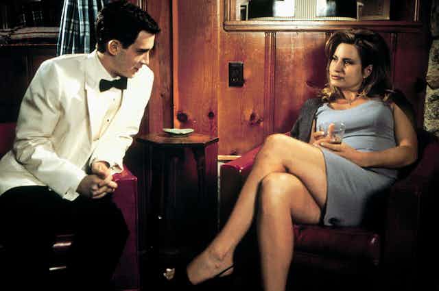Finch, a teen boy, sits in a white tuxedo. Opposite him, Stifler's Mom wears a blue dress and sips whiskey.