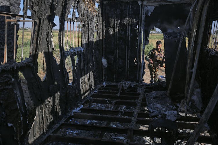 A soldier in camouflage steps into a destroyed vehicle that appears charred from the inside.