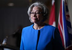 A grey-haired woman with glasses in a blue suit smiles faintly.