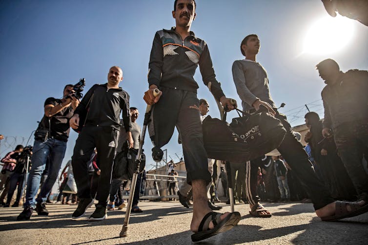 A group of men, one with crutches and an amputated leg, walk, followed by some men with cameras photographing them.