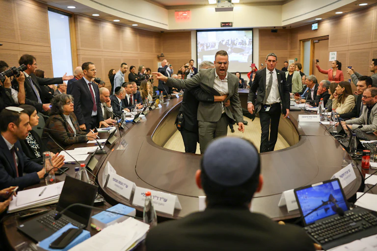A meeting of people sitting and standing around a horseshoe-shaped desk, with an angry man in the middle being pulled back.