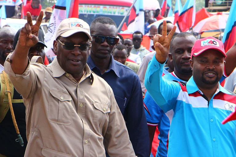 Two man holding up their right hands in a two-finger salute - there is a crowd of other men behind them.