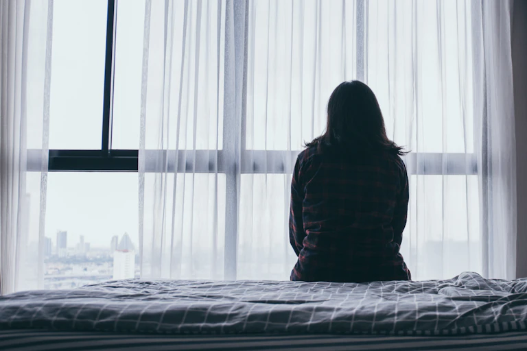 a lonely woman sits one a bed, silhouetted as she looks out of a window
