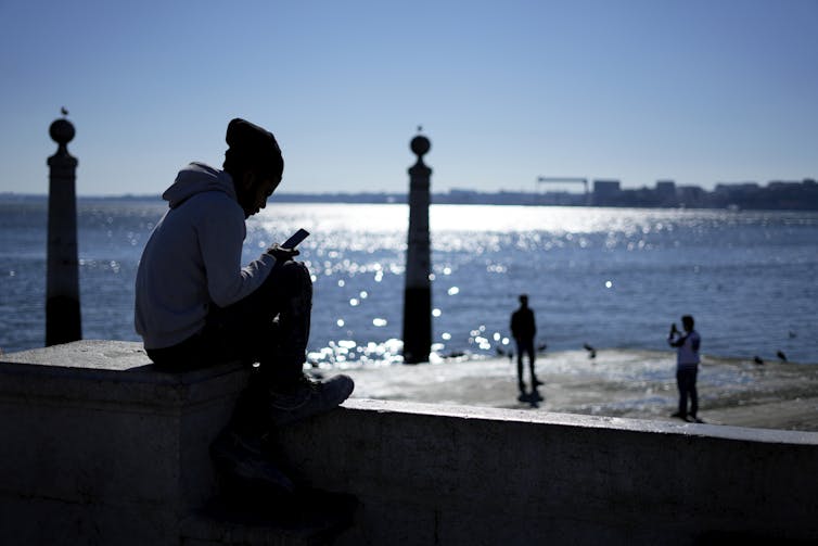 A teenager looks at his phone with shimmering water behind him and people walking along a pier.