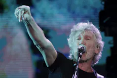 A man with grey hair and a beard sings into a microphone with one arm raised.