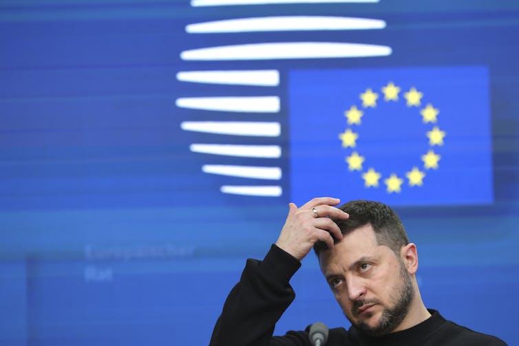 A bearded man with dark hair with his hand on his head standing in front of a European Union flag.