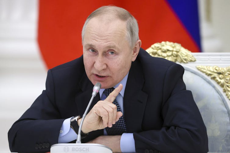 A balding man raises his finger while speaking into a microphone. A red and blue flag is behind him.