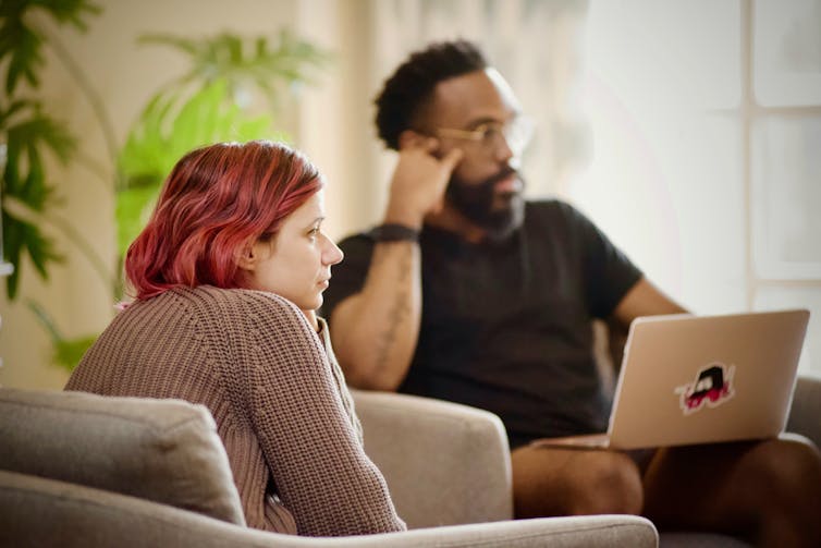 A white woman with pink hair and a Black man listen to someone off-camera. The man has a laptop on his lap.