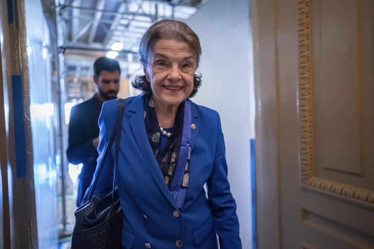 An older woman in a bright blue double-breasted jacket, her hair pulled back, smiling and walking down a hallway.