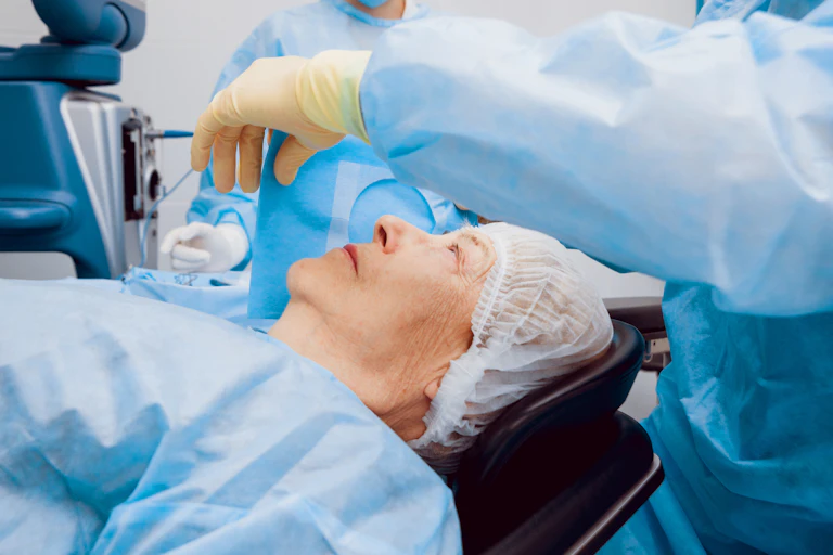 An aging woman lies on an operating table with doctors in scrubs surrounding her.
