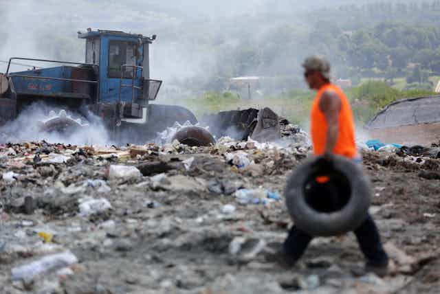 A tractor drives over trash as a man walks past carrying a tire.