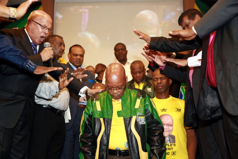 A man kneeling with his eyes closed as others hold out their hands over his head and one man speaks into a microphone