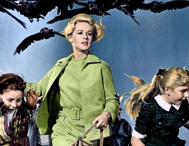 A blonde woman in a pea green coat runs as she is chased by a flock of crows