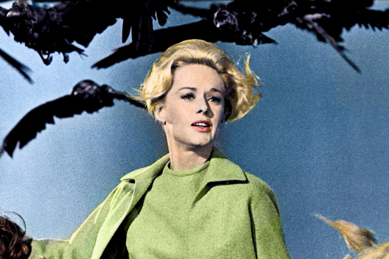 A blonde woman in a pea green coat runs as she is chased by a flock of crows