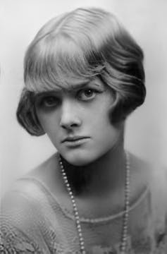 A black and white portrait of a young Daphne du Maurier with a bob and pearl necklace.