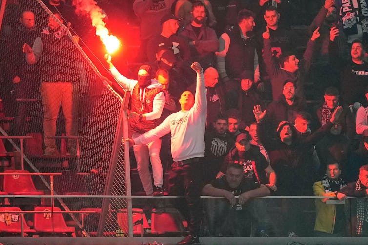 A man sets off a firework at a soccer match while another supporter makes a clenched fist salute.