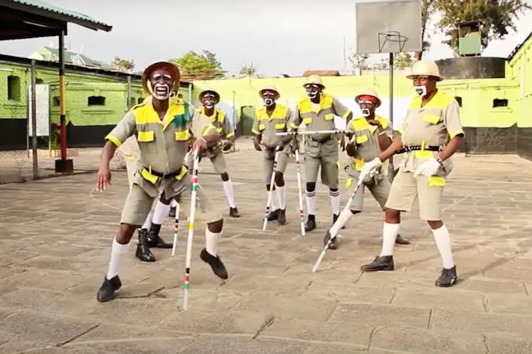 A troupe of men hold sticks. They are dressed comically in army uniforms with padded bellies and exaggerated beards and moustaches. They are in a courtyard.