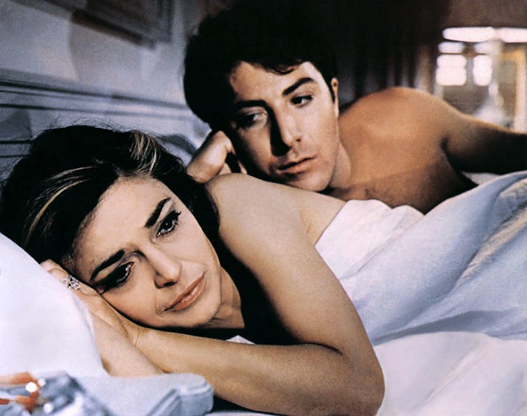 Dustin Hoffman and Anne Bancroft lie in bed, post coital. She is turned away from him and he is behind her, looking searchingly at her face.