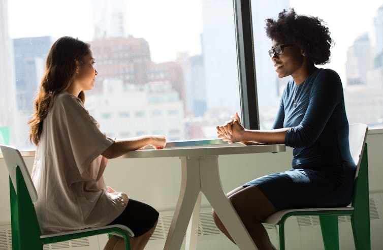 Two women sit at a desk beside a window. One speaks while the other listens.