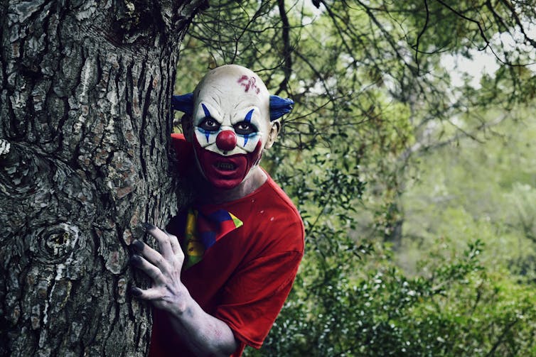 Un clown effrayant sort de derrière un arbre. Il porte un haut rouge. Il est maquillé en blanc et affiche un large sourire sinistre peint en rouge