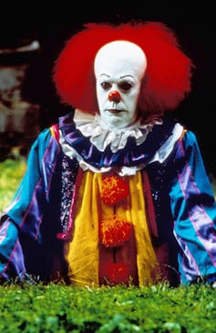 The head and torso of a creepy clown appear through a field. He has a long white face with a shock of red hair. He is wearing a clown suit with a ruffled neck and pompoms down the front.