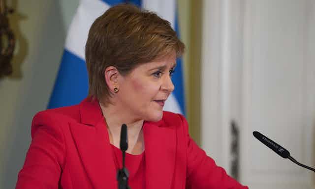 Close up photo of Nicola Sturgeon in a red suit, delivering her resignation speech from a podium in front of a Scottish flag