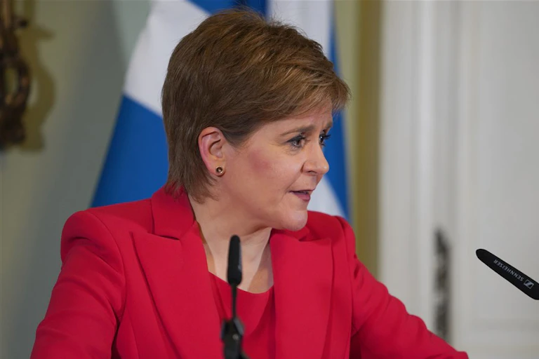 Close up photo of Nicola Sturgeon in a red suit, delivering her resignation speech from a podium in front of a Scottish flag