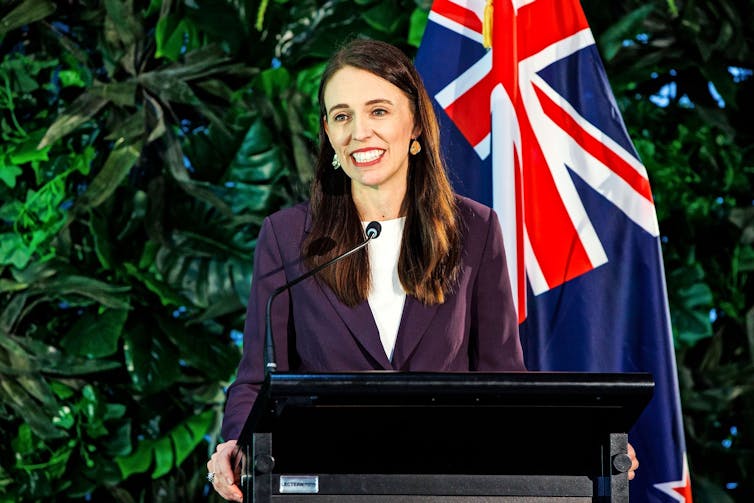 Jacinda Ardern stands at a podium in front of a wall of foliage and a New Zealand flag.