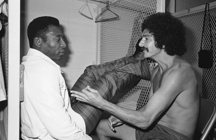 A shirtless soccer player talks with Pelé in white jacket, reclining.