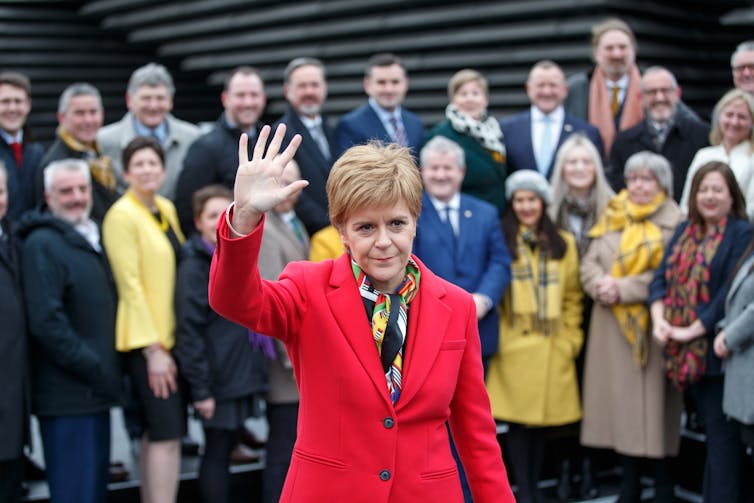 Nicola Sturgeon waves while wearing a red suit and standing in front of a group of SNP MSPs