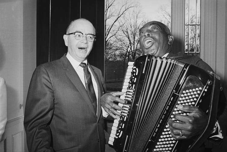 A white man dressed in a business suit is singing along with an elderly black man who is playing an accordion.