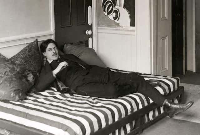 Wyndham Lewis wears a suit and boots, lying on a mattress in on the floor of his home. In one hand he holds a cigarette.