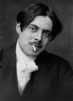 Wyndham Lewis has longish hair parted curtains-style in the middle. He smokes a cigarette under his neatly manicured moustache.