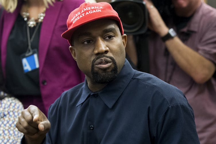 Kanye West in a press conference wearing a blue button down shirt and a red 'Make America Great Again' cap.
