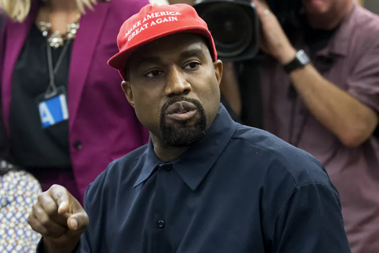 Kanye West in a press conference wearing a blue button down shirt and a red 'Make America Great Again' cap.